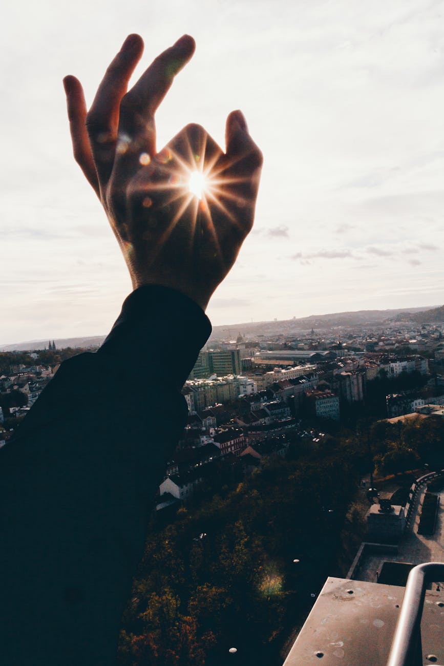 A person holding their hand to make an Okay sign with the sun in the center while overlooking the city. 